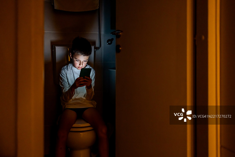 年轻男孩在昏暗的浴室里坐在马桶上使用智能手机图片素材