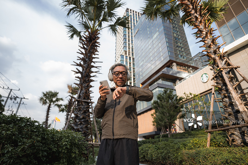 老年男子在跑步后休息，查看他的智能手表和智能手机上的活动追踪应用。图片素材