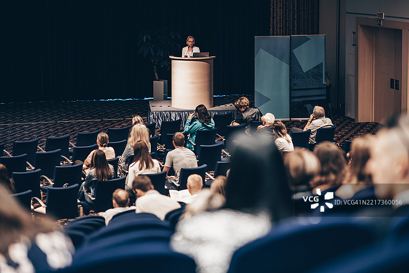 女性演讲者在企业商务会议上发言，观众中有不明身份的人士。图片素材