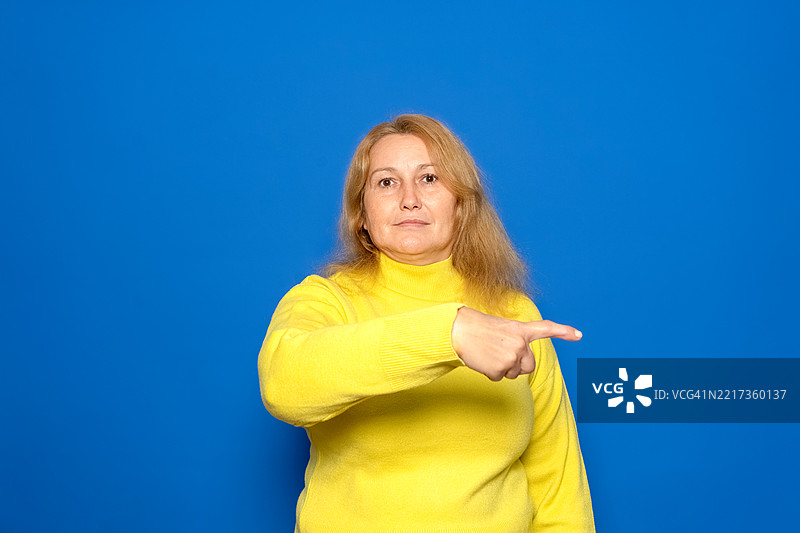 一位四十多岁的白人女性穿着黄色毛衣，害羞地指着。图片素材