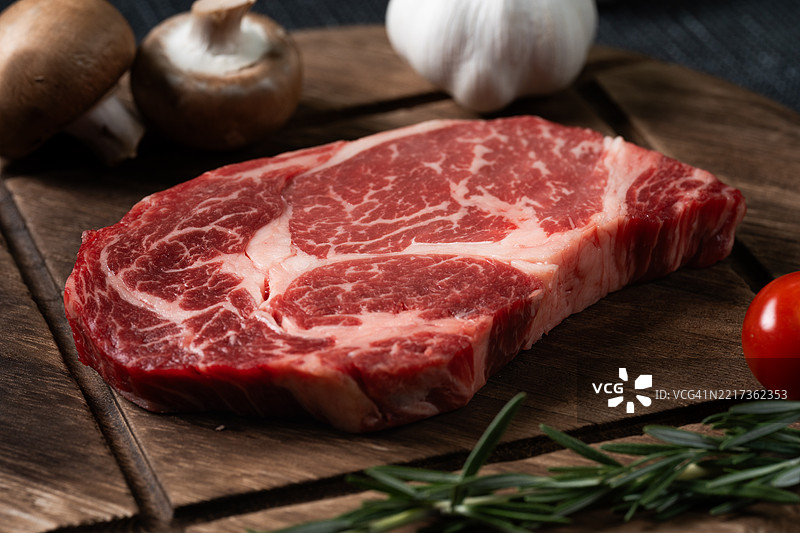 新鲜的牛排上放着迷迭香、西红柿、蘑菇和大蒜，摆放在木质盘子上。图片素材