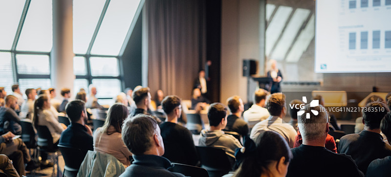 在商业活动的会议厅中，演讲者正在发言，背面是一些无法识别的人群。图片素材
