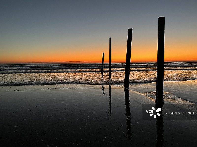 海滩上，木柱在黎明时分映衬着发光的橙色天空。图片素材