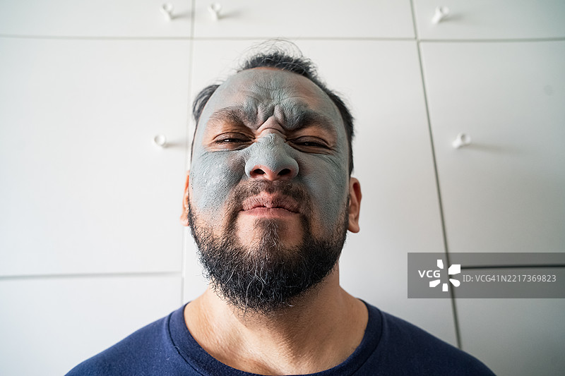 兴奋的成熟亚洲男性的美容肖像，独自站在白色背景前，脸上涂着美容泥膜。图片素材