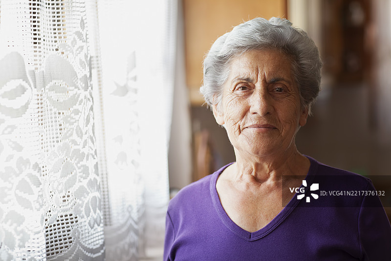 微笑的老年女性在窗边，窗帘上有蕾丝花边图片素材