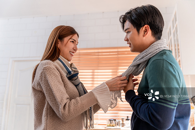 亚洲年轻夫妇在冬季的家中享受温馨时光。迷人的妻子为丈夫系上温暖的围巾，感到快乐和放松，在家中的厨房里舒适地度过时光。图片素材