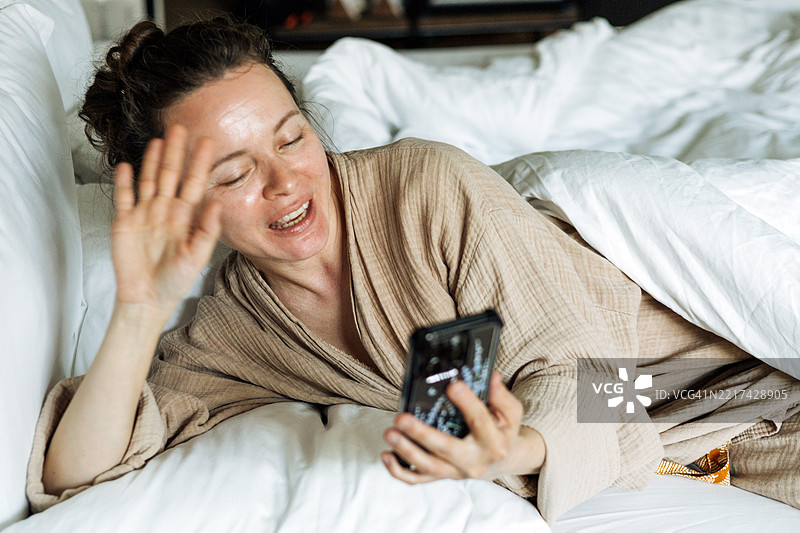 女人在自然光环境中躺在床上微笑并挥手进行视频通话。图片素材