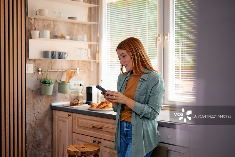 年轻女性在厨房里吃早餐时使用智能手机图片素材