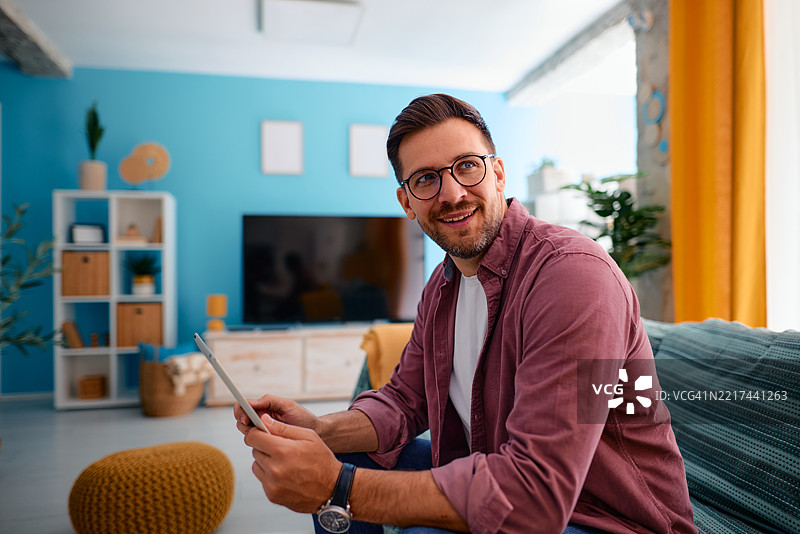 年轻男子在客厅使用平板电脑，微笑着望向一旁。图片素材