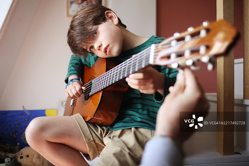一个小男孩在他的卧室里弹吉他，我们可以看到他父亲的手在调音。图片素材