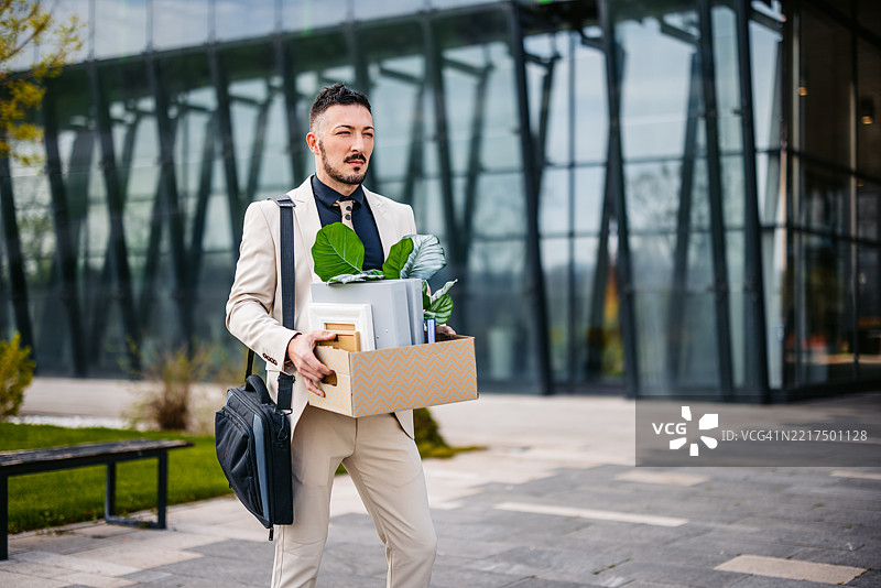 悲伤的中年商人手里拿着办公桌上的物品，站在一座企业大楼前。图片素材