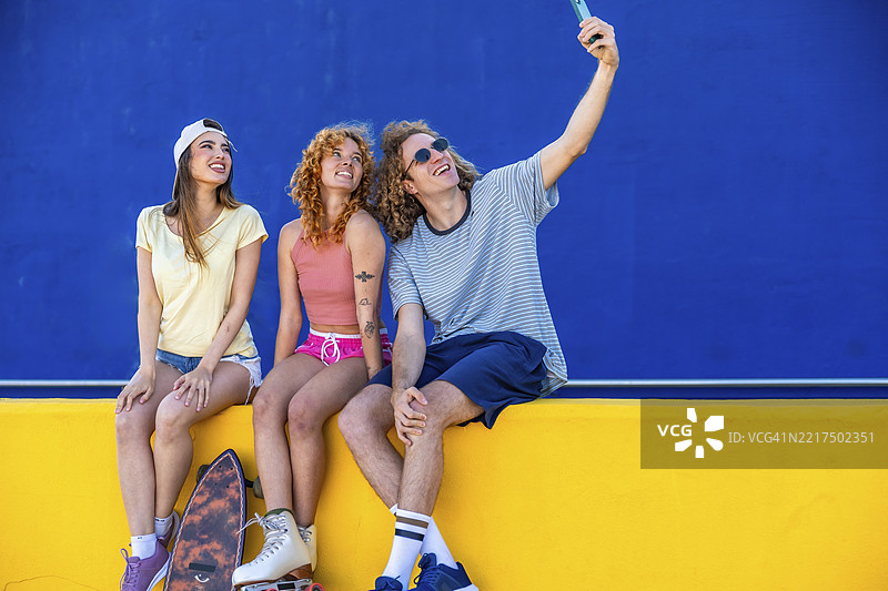 三个快乐的朋友在蓝色背景的黄色墙壁前用智能手机自拍图片素材