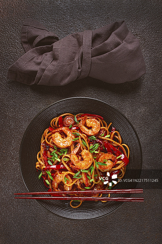 黑碗中的乌冬炒虾面，配有红椒和青葱，旁边放着筷子，背景为深色。图片素材