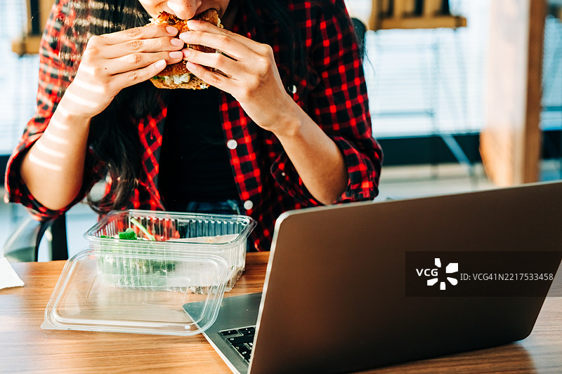 女人坐在桌子前吃三明治，盯着她的笔记本电脑。图片素材