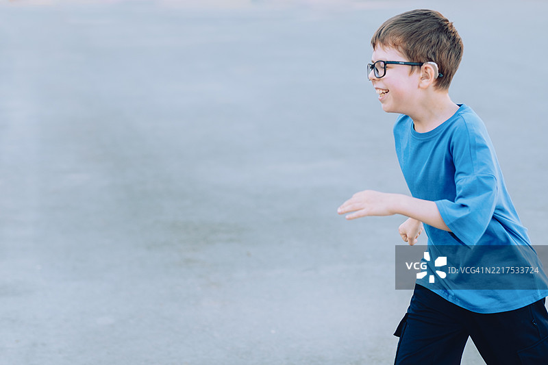 11岁男孩戴着阅读眼镜和助听器，在游戏中奔跑，穿着蓝色T恤，背景是沥青。图片素材