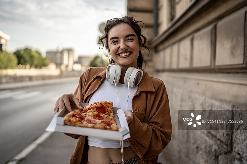 微笑的女性在户外手持披萨盒，脖子上挂着耳机图片素材