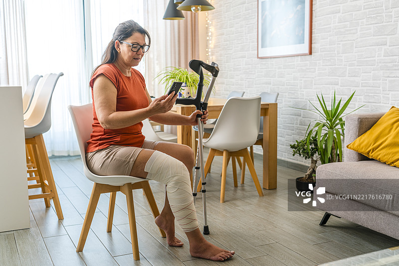 坐在椅子上的女性，腿部包扎着，手持拐杖，正在用手机与医生或保险公司进行视频通话。图片素材