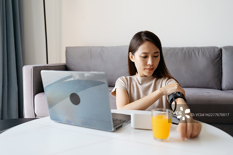 亚洲华裔女性在家中使用笔记本电脑并用血压计测量自己的血压图片素材