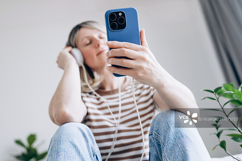 年轻的金发女性在舒适的家中，戴着耳机享受来自智能手机的音乐。图片素材