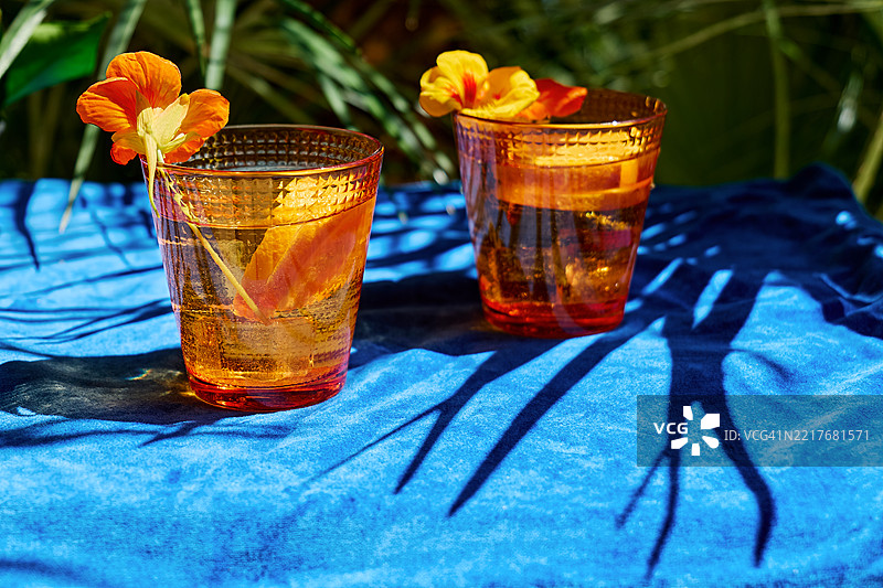 热带花园中，蓝色桌布上摆放着冰镇的花草水，里面有可食用的金莲花和橙子片。图片素材