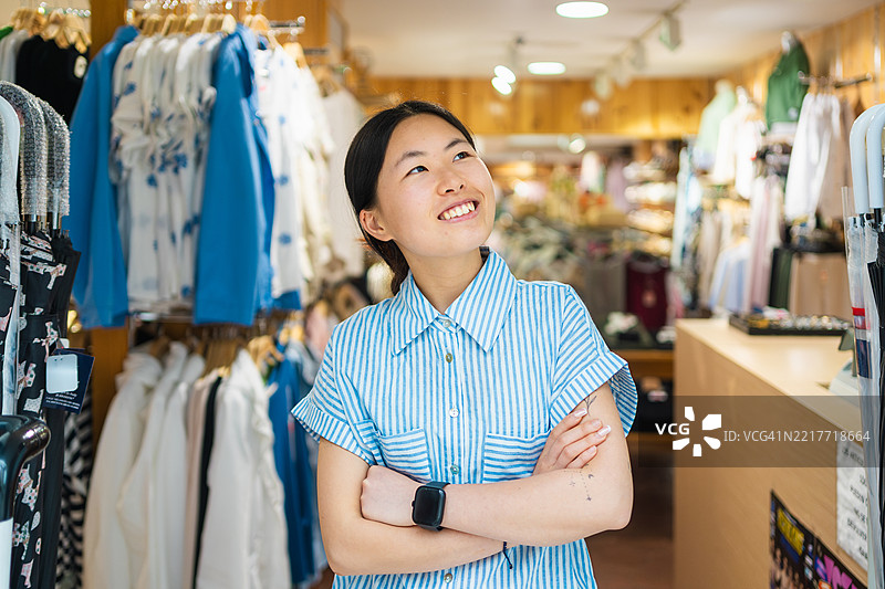 年轻女销售助理微笑着仰望，双臂交叉站在服装店内图片素材