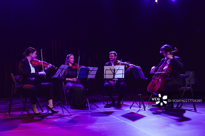 四位弦乐四重奏音乐家在剧院舞台上并肩演奏古典音乐，演奏乐器包括中提琴、大提琴和小提琴。图片素材