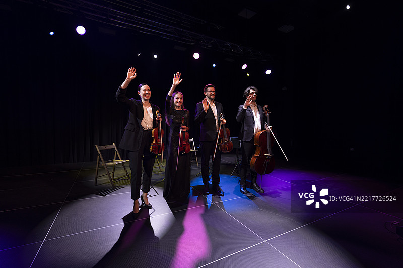弦乐四重奏的音乐家们在舞台上排成一排，举手感谢观众，表演结束时灯光投射在他们身上。图片素材