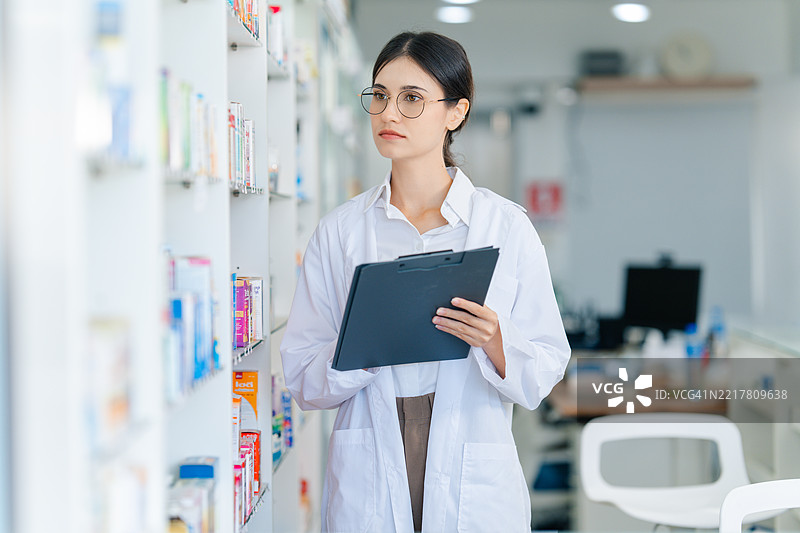药剂师在现代药房检查货架并在夹板上记录库存。库存管理、药物控制和专业医疗服务的概念。图片素材