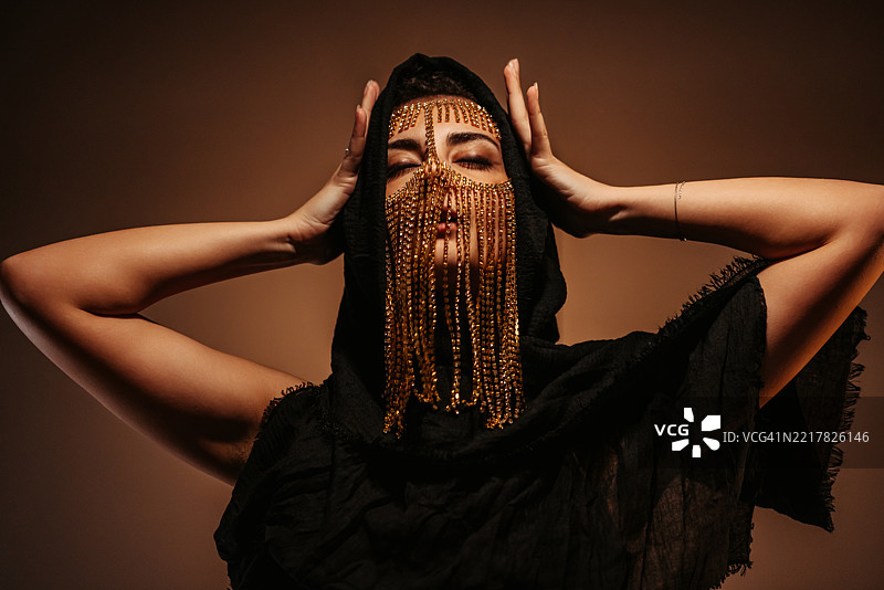 年轻女性佩戴中东风格面具，黑色兜帽在金色霓虹灯下闪耀图片素材