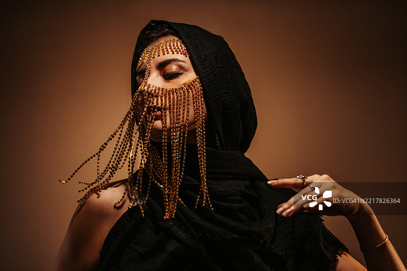 年轻女性佩戴中东风格面具，黑色兜帽在金色霓虹灯下。图片素材