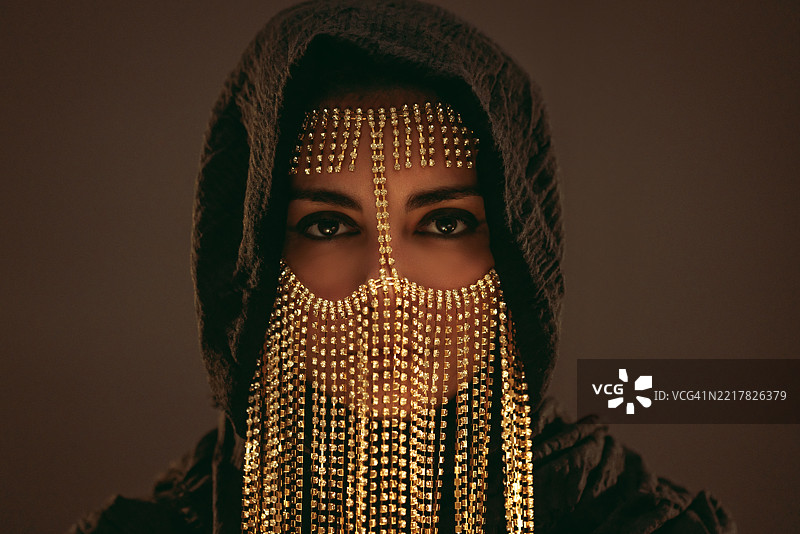 年轻女性佩戴中东风格面具，黑色兜帽在金色霓虹灯下闪耀图片素材