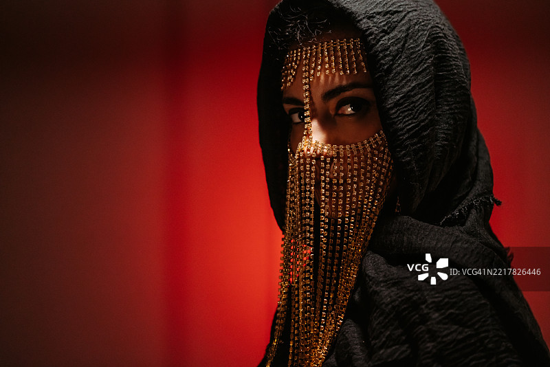 年轻女性佩戴中东风格面具，黑色兜帽在红色霓虹灯下照耀图片素材