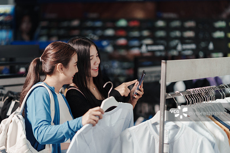 两位亚洲女性在购物，查看智能手机上的产品详情，选择运动服，数字生活方式，现代零售购物，友谊，健身时尚。图片素材