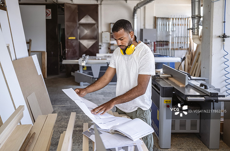专注的多种族男性木匠在明亮的工作室中查看图纸，周围有现代工具和木材材料图片素材