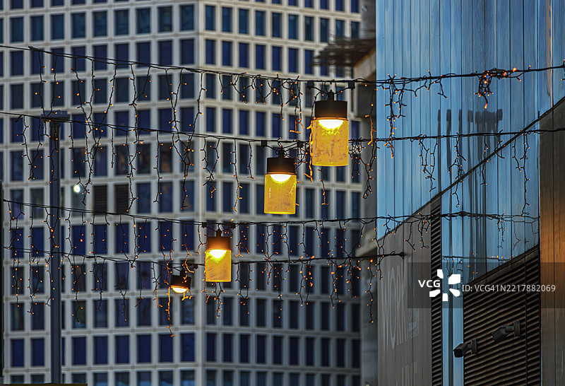 温暖的悬挂灯和串灯在现代玻璃和格子图案建筑前闪耀。图片素材