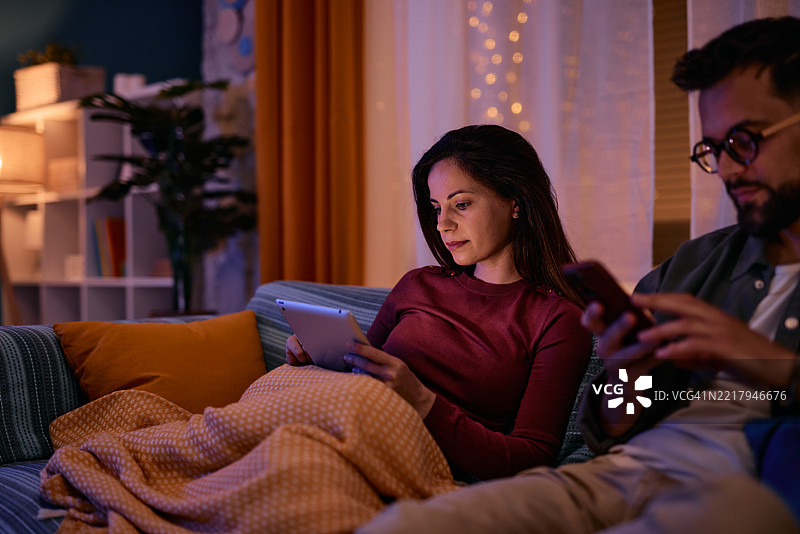 情侣在晚上舒适地坐在沙发上使用平板电脑和智能手机。图片素材