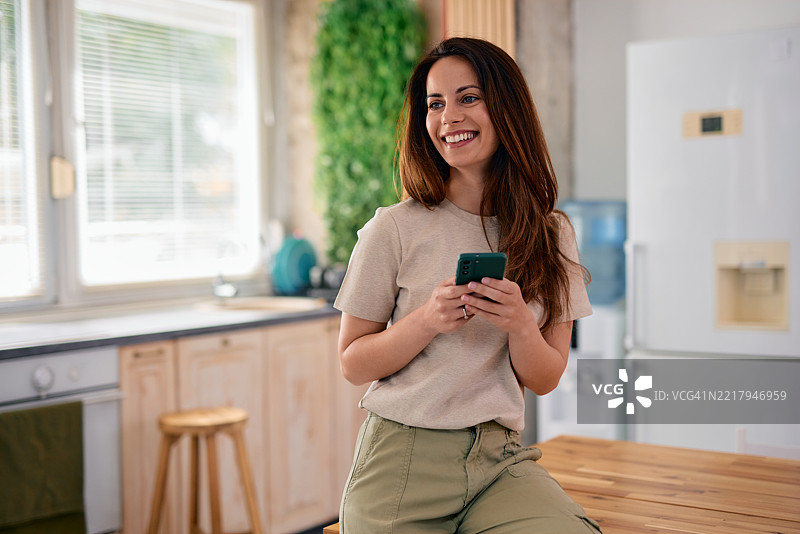 年轻女性在现代厨房中使用智能手机并微笑图片素材