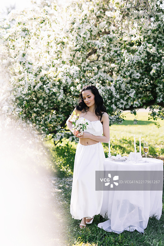 一位多种族的美丽迷人的年轻女性穿着优雅的白色服装，享受大自然和盛开的植物与花朵。一位新娘穿着婚礼服装，站在公园草地上装饰好的桌子旁。图片素材