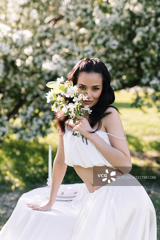 一位多种族的美丽迷人的年轻女性穿着优雅的白色服装，享受大自然和盛开的植物与花朵。一位新娘穿着婚礼服装，站在公园草地上装饰好的桌子旁。图片素材
