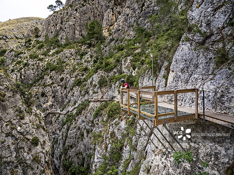 一位女性在玻璃底观景平台上休息，沿着嵌入峡谷岩壁的木栈道行走。雷尤桥（Pasarela de Relleu）图片素材