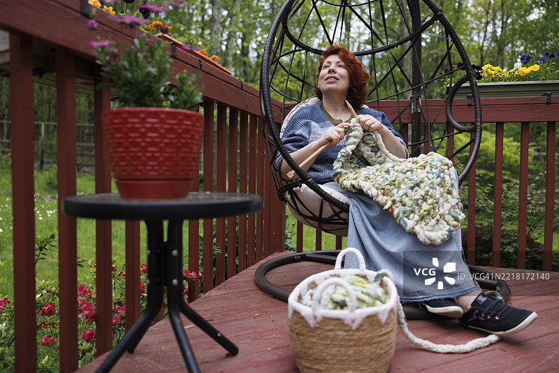 篮子里装着用品，红发女子正在进行阿富汗毯项目。白人女性坐在木甲板上的球形椅子上，享受她的爱好。图片素材