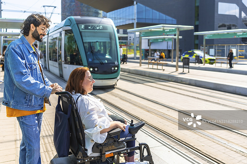 一名男子在现代车站推着一位微笑的女性坐在电动轮椅上等待电车。图片素材