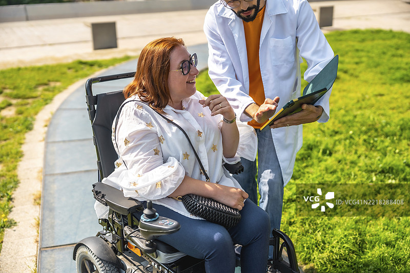 医生在公园里向坐在轮椅上的微笑女性展示医疗信息，宣传无障碍设施和医疗支持图片素材