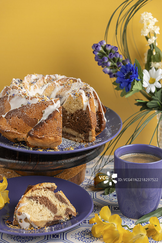 切好的大理石圆环蛋糕，放在蓝色盘子上，淋上糖霜，周围有花朵和一杯咖啡，背景为黄色。图片素材
