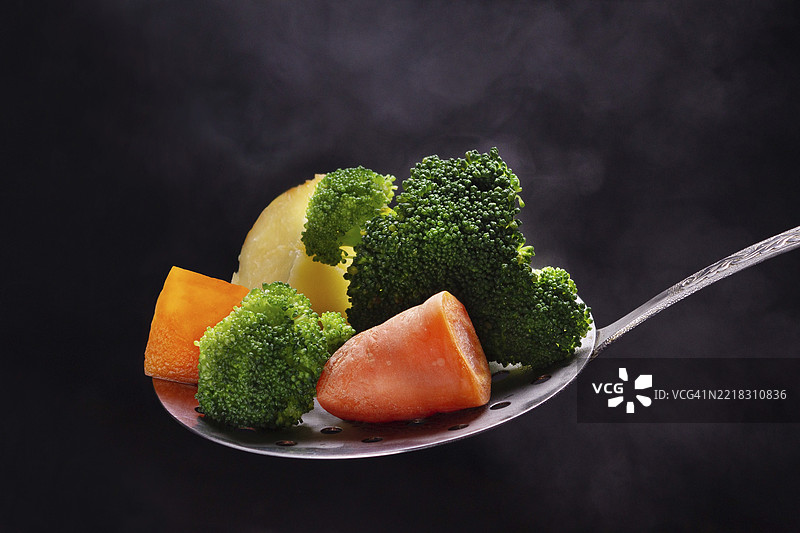 蒸汽中，带有孔的勺子上放着西兰花、胡萝卜和土豆，焦点选择性突出。图片素材