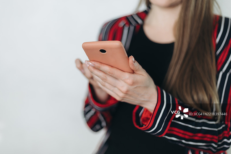 一位穿着红黑色衣服的年轻女性在白色墙壁前使用智能手机，面容模糊不清。图片素材