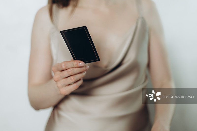 一位模糊不清的女性穿着浅色连衣裙，手持一张黑色扑克牌，站在白色墙壁旁。图片素材