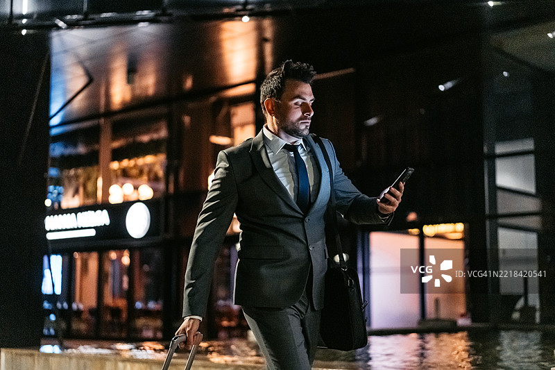 中年商人在夜晚走在公司大楼前，边走边用手机，身旁推着行李箱。图片素材