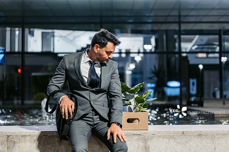 伤心的中年商人坐在公司大楼前，手里拿着一箱办公用品。图片素材