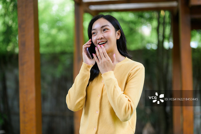 年轻的亚洲女性在大自然和树木的环绕中，开心地微笑和欢笑，接收到令人兴奋的手机消息。图片素材